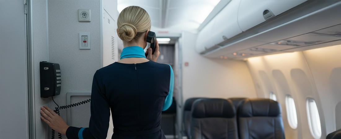 Azafata (TCP) de pie en el pasillo del avión, con uniforme azul y sombrero, vista desde atrás mientras mira hacia el fondo de la cabina.